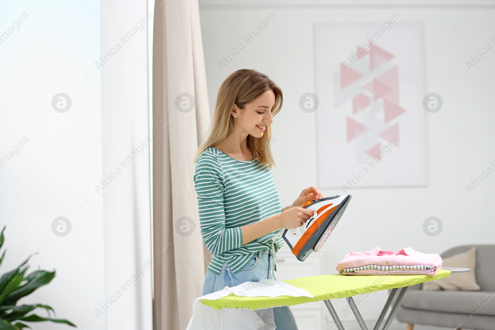 Photo of Young pretty woman ironing clean laundry on board indoors