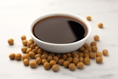 Photo of Soy sauce in bowl and soybeans on white table, closeup