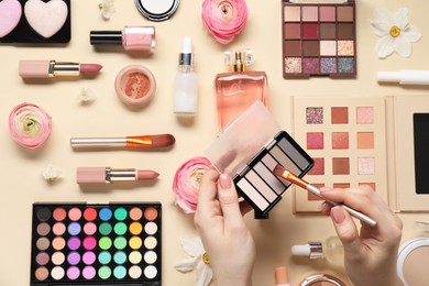 Photo of Woman applying eye shadow palette on brush, closeup. Different makeup products and beautiful spring flowers on beige background, top view