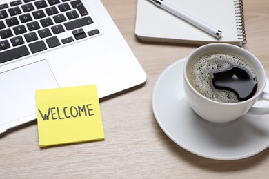 Paper note with word Welcome, laptop and cup of coffee on wooden table