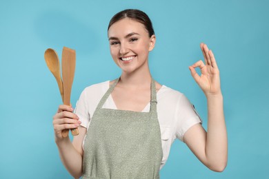 Beautiful young woman in clean apron with kitchen tools on light blue background