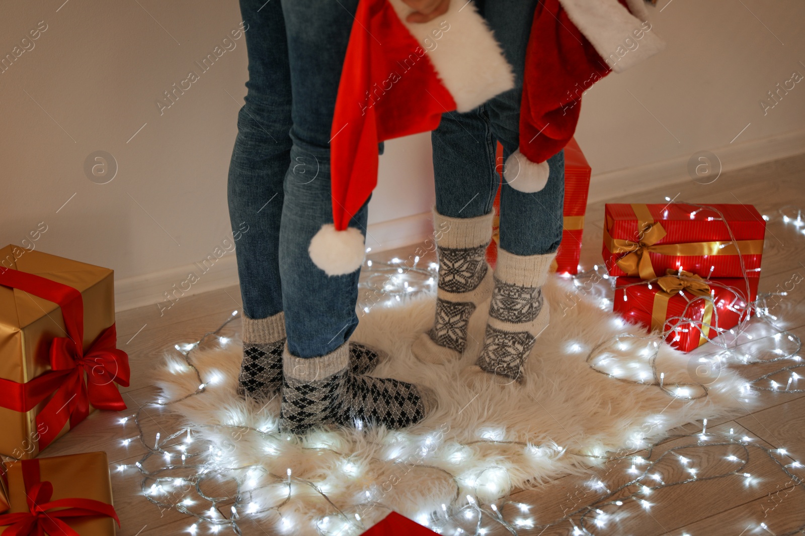 Photo of Young couple with Santa hats standing on rug near Christmas lights and gift boxes, closeup