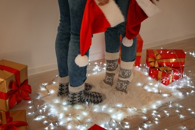 Photo of Young couple with Santa hats standing on rug near Christmas lights and gift boxes, closeup