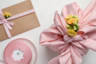 Furoshiki technique. Gift packed in pink fabric, card, flowers and ribbon on white table, flat lay