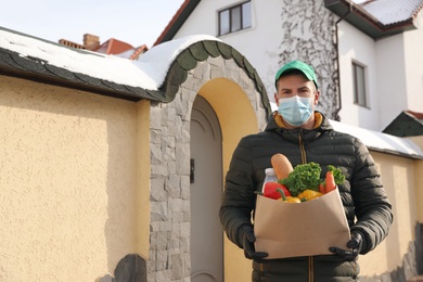 Courier in medical mask holding paper bag with groceries near house outdoors. Delivery service during quarantine due to Covid-19 outbreak