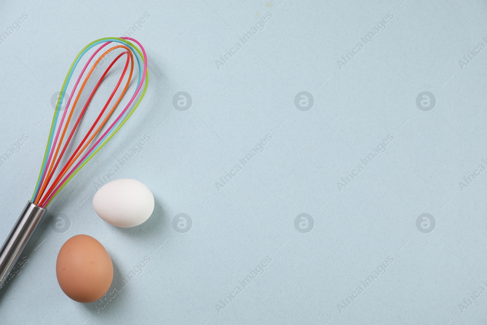 Photo of Colorful whisk and raw eggs on light blue background, flat lay. Space for text