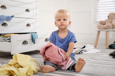 Photo of Cute little boy playing with clothes near dresser on floor at home