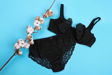 Photo of Elegant black plus size women's underwear and cotton branch with fluffy flowers on light blue background, flat lay