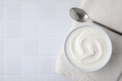 Photo of Delicious natural yogurt in bowl and spoon on white tiled table, top view. Space for text