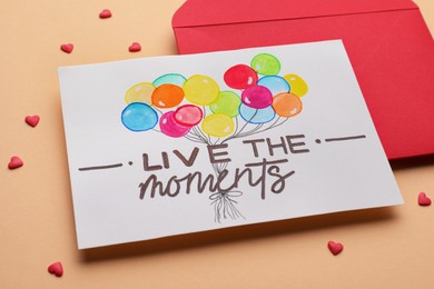 Photo of Card with phrase Live The Moments and red envelope on beige background, closeup