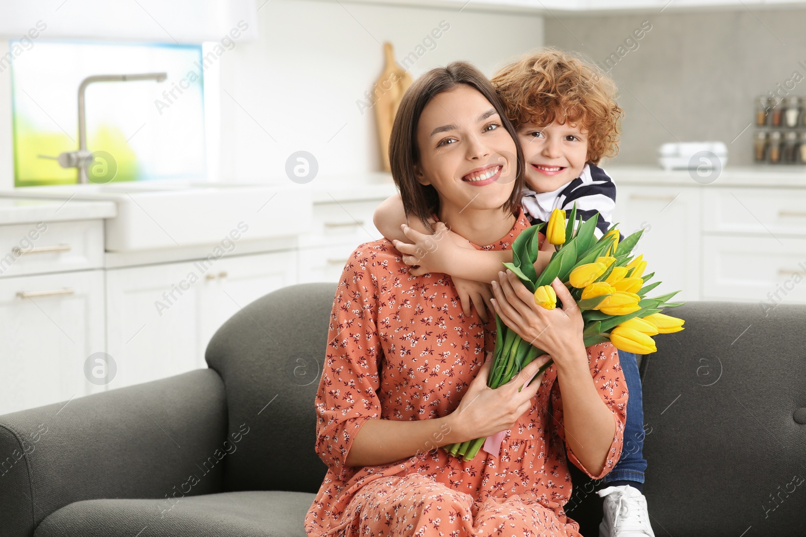 Photo of Little son congratulating his mom with Mother`s day at home. Woman holding bouquet of yellow tulips