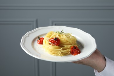 Photo of Waiter holding plate of tasty capellini with tomatoes and cheese near grey wall, closeup. Exquisite presentation of pasta dish