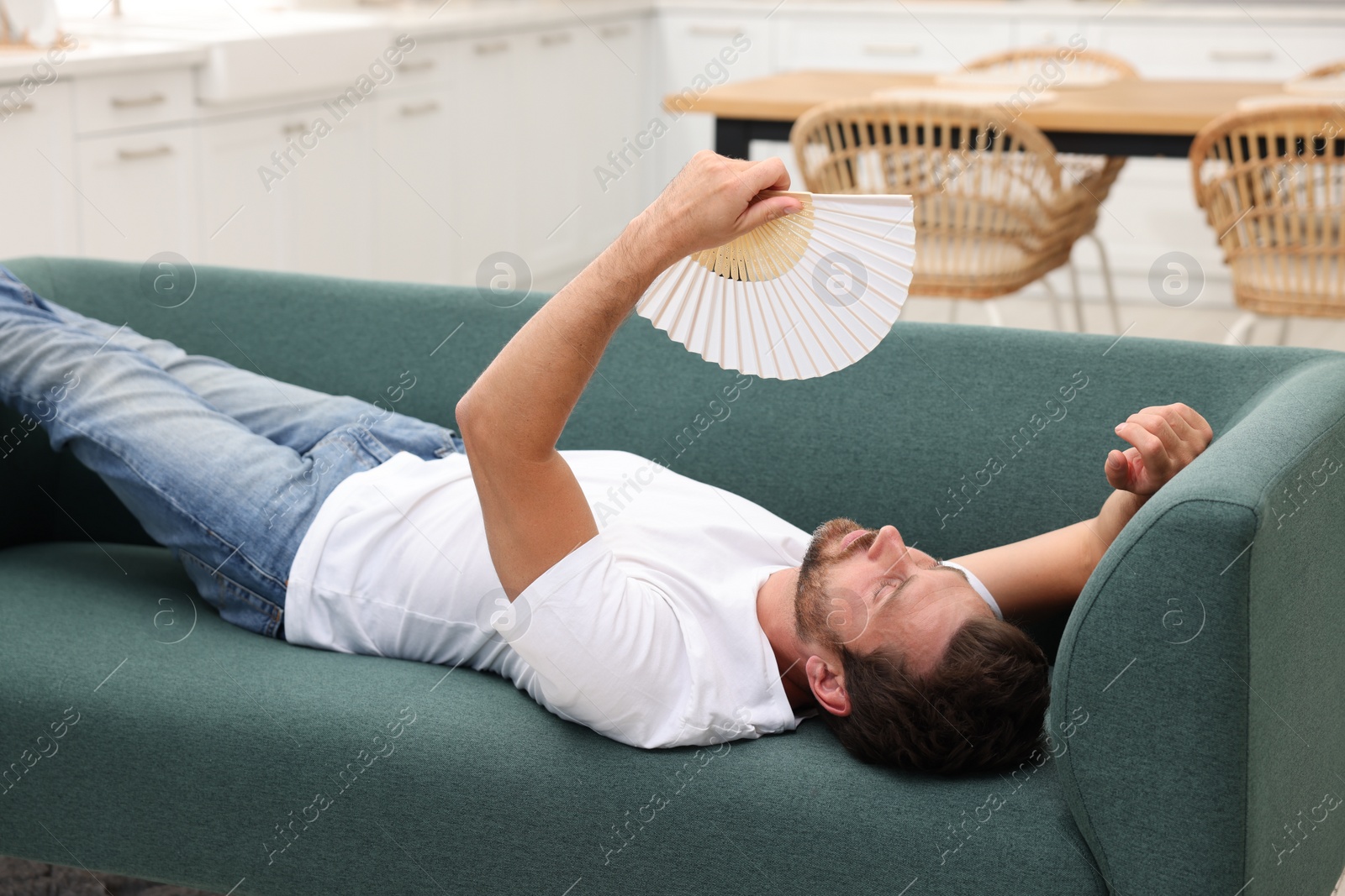 Photo of Man waving white hand fan to cool himself on sofa at home