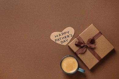 Photo of Heart shaped card with phrase HAPPY FATHER'S DAY, gift box and cup of aromatic coffee on brown background, flat lay. Space for text