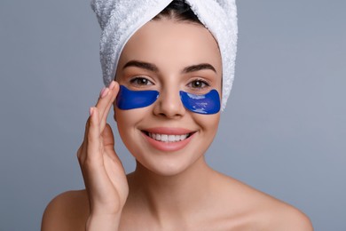 Beautiful young woman with under eye patches and hair wrapped in towel on grey background