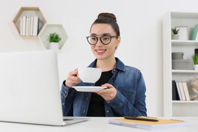 Home workplace. Happy woman with cup of hot drink looking at laptop at white desk in room