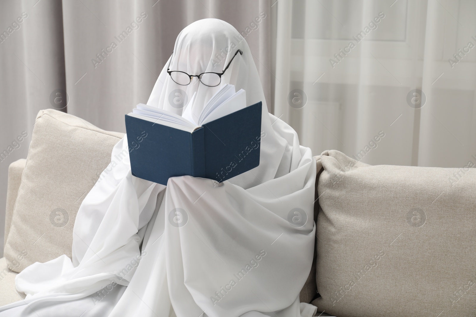 Photo of Creepy ghost. Person covered with white sheet reading book on sofa at home