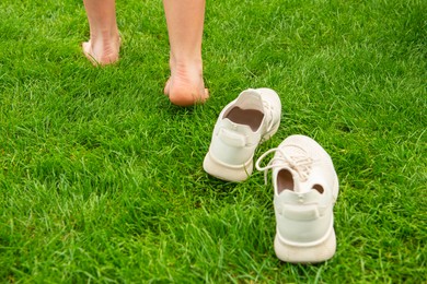 Photo of Woman leaving her sneakers and walking away barefoot on green grass, closeup