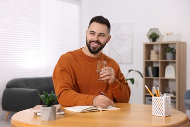 Photo of Young man with glass of water writing in notebook at wooden table indoors