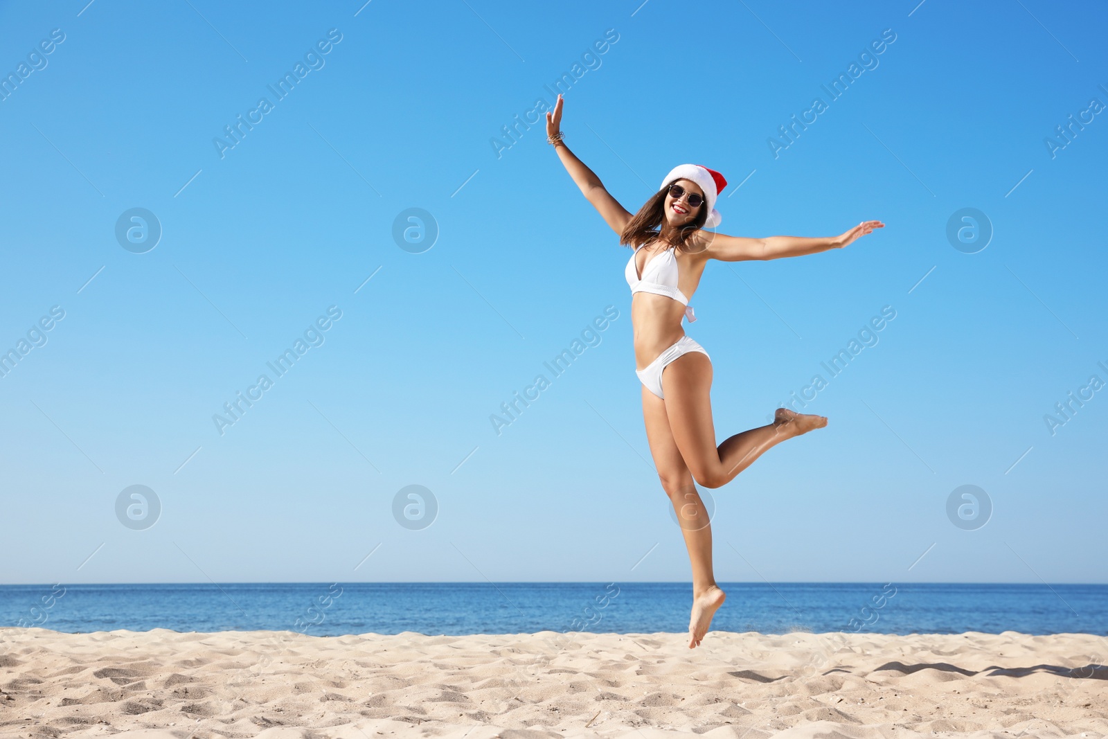 Photo of Young woman in Santa hat and bikini jumping on beach, space for text. Christmas vacation