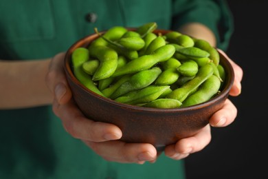 Woman holding bowl with green edamame beans in pods on black background, closeup