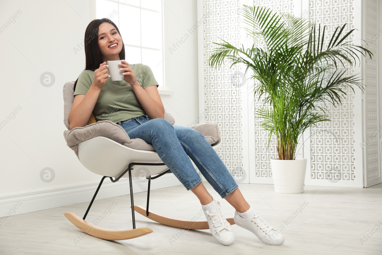 Photo of Young woman with cup of drink relaxing in rocking  chair at home