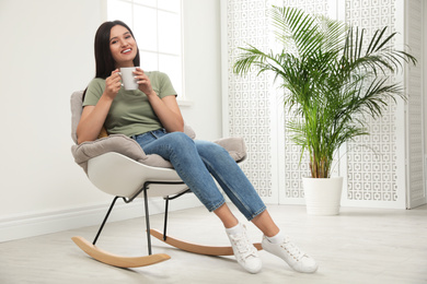 Young woman with cup of drink relaxing in rocking  chair at home
