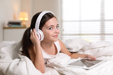 Photo of Cute little girl with headphones and tablet listening to audiobook in bed at home