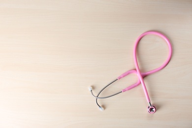 Pink stethoscope as awareness ribbon on wooden background, top view with space for text. Breast cancer concept