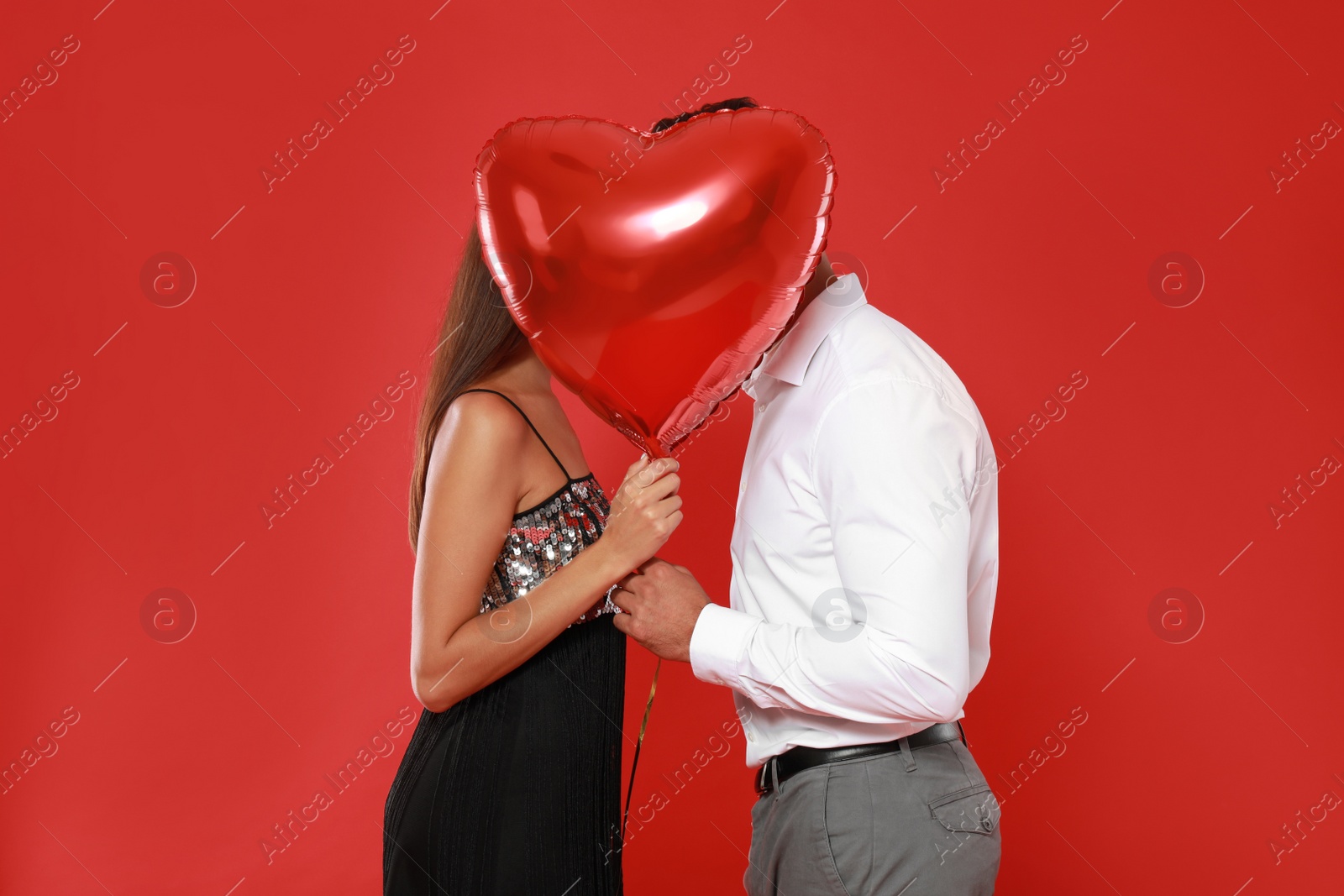 Photo of Couple kissing while hiding behind heart shaped balloon on red background