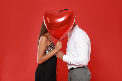 Photo of Couple kissing while hiding behind heart shaped balloon on red background