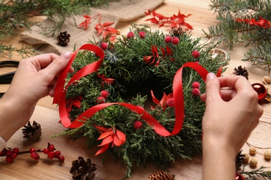 Photo of Florist making beautiful Christmas wreath with berries and red ribbon at wooden table, closeup
