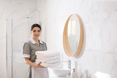 Photo of Young chambermaid holding stack of fresh towels in bathroom