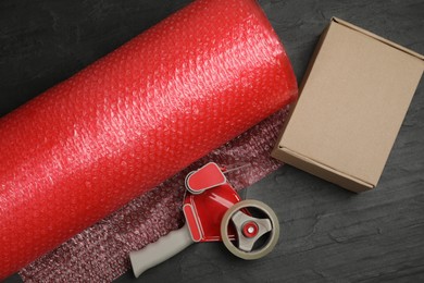 Photo of Bubble wrap roll, cardboard box and tape dispenser on black background, flat lay