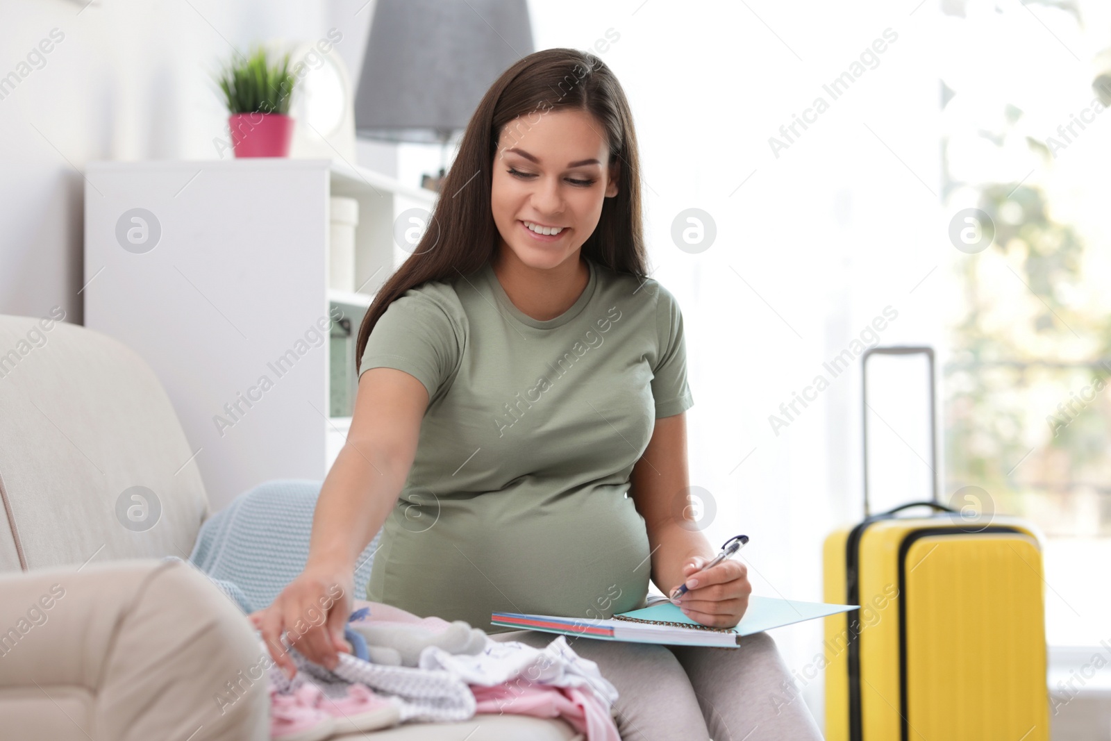 Photo of Pregnant woman making list while packing suitcase for maternity hospital in living room