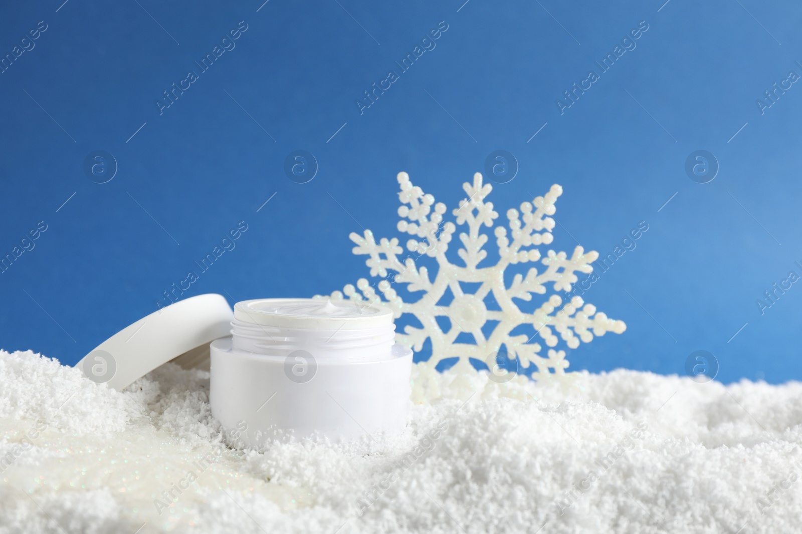 Photo of Jar of hand cream and decorative snowflake on snow against blue background. Winter skin care