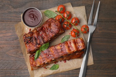 Photo of Tasty roasted pork ribs served with sauce, basil and tomatoes on wooden table, top view