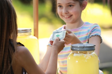 Photo of Little girl selling natural lemonade to kid in park. Summer refreshing drink