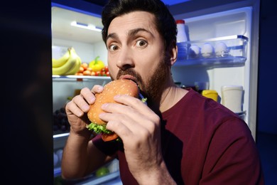 Photo of Man eating burger near refrigerator in kitchen at night. Bad habit