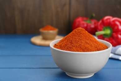 Photo of Bowl with aromatic paprika powder and fresh bell peppers on blue wooden table. Space for text