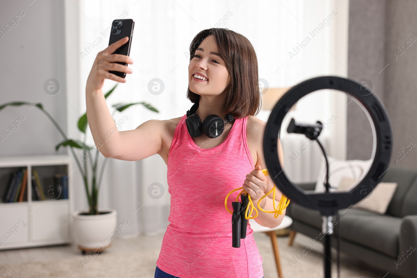 Photo of Happy sports blogger holding skipping rope while streaming online fitness lesson with smartphone at home