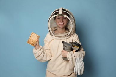 Photo of Beekeeper in uniform holding smokepot and hive frame with honeycomb on light blue background