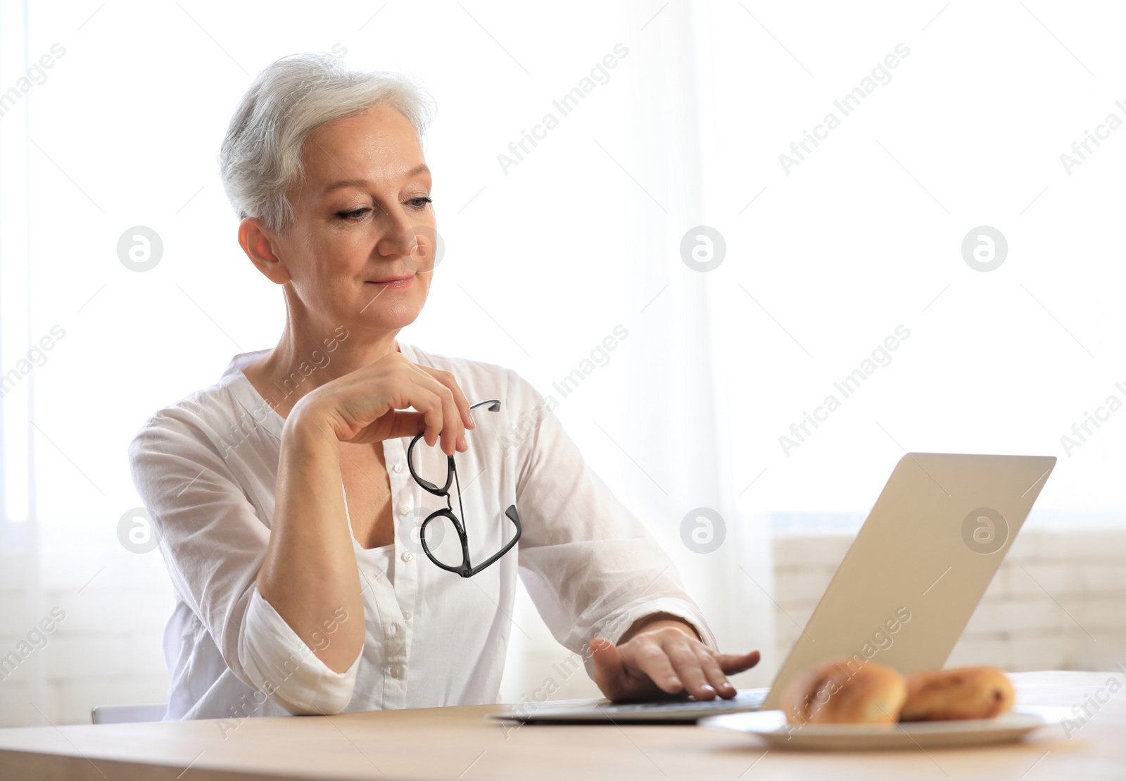 Photo of Mature woman with laptop sitting at table in kitchen. Smart aging