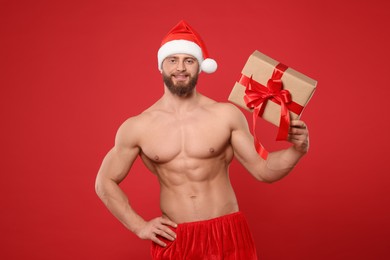 Attractive young man with muscular body in Santa hat holding Christmas gift box on red background