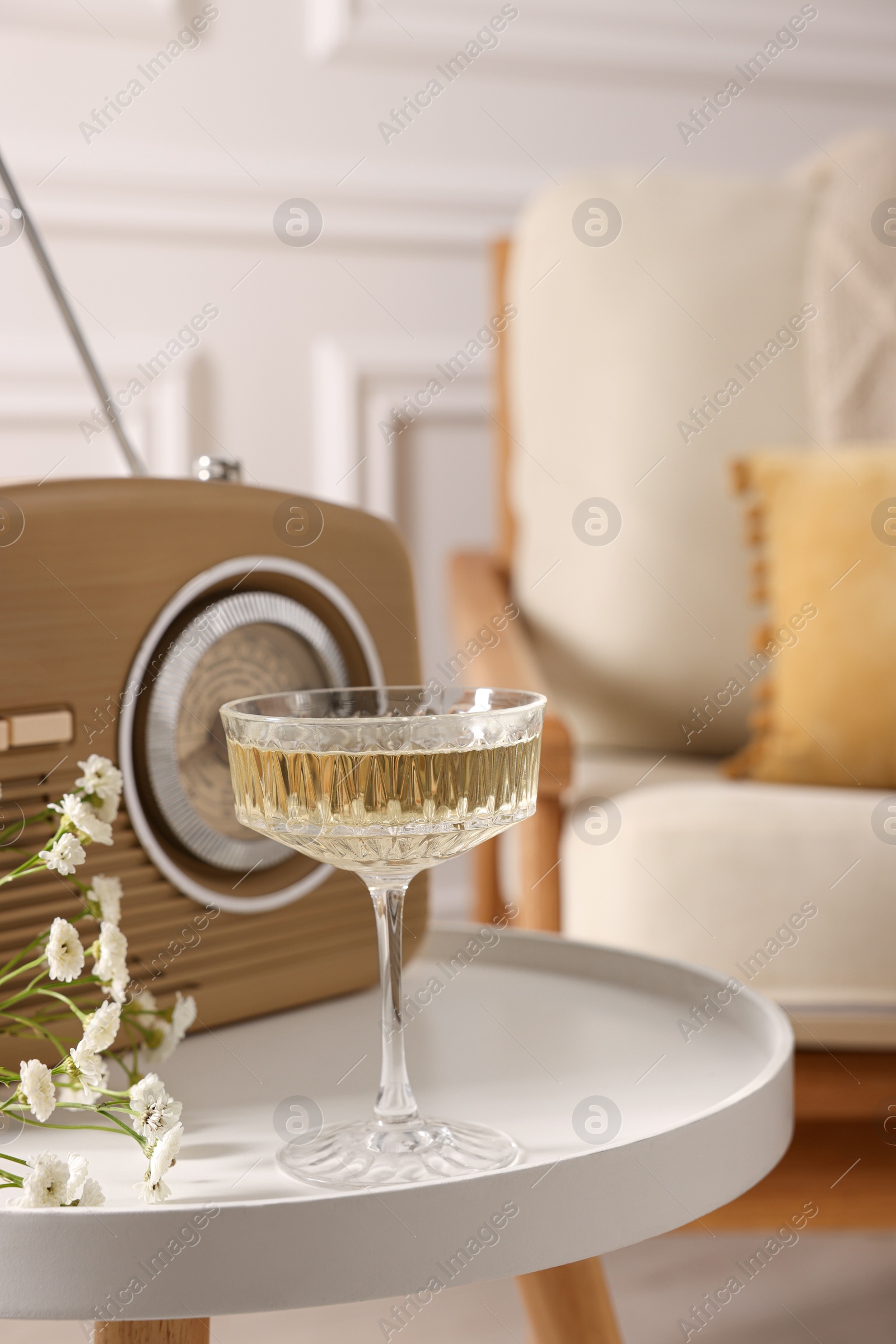 Photo of Glass of alcohol drink, flowers and radio set on table in room, closeup. Relax at home