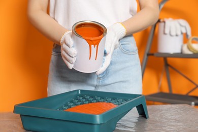 Photo of Woman pouring orange paint from can into tray at table, closeup