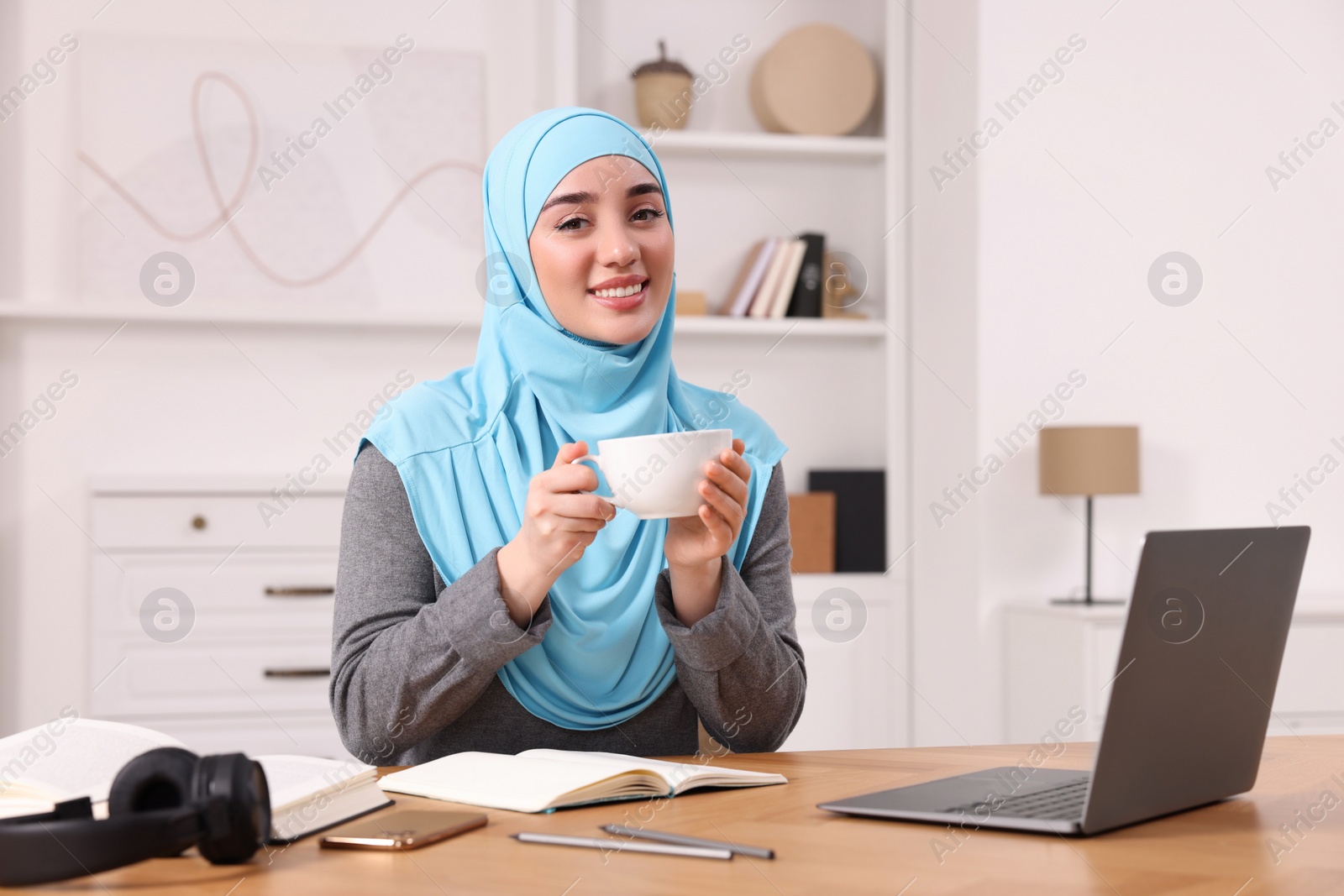 Photo of Muslim woman in hijab with cup of coffee using laptop at wooden table in room