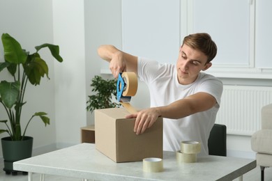 Young man packing box with adhesive tape indoors