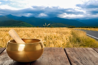 Image of Golden singing bowl and mallet on wooden surface in wheat field, space for text