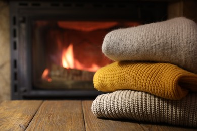 Stack of sweaters on wooden table near fireplace at home, closeup. Space for text
